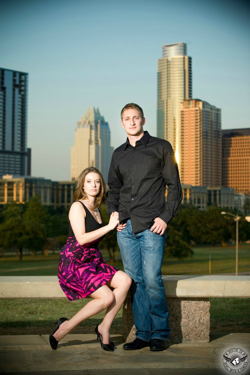attractive couple in red skirt an black tank top and black button down shirt with blue jeans at buttler park on Doug Sam hill summit with the skyline of the city of austin in  the back ground with a blue sky and the frost bank building visible